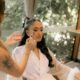 A bride in a white robe getting makeup applied by an artist, surrounded by natural light.