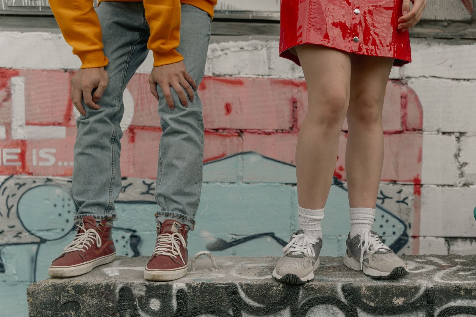 Two people in stylish streetwear against a graffiti backdrop, highlighting urban fashion with jeans and sneakers.