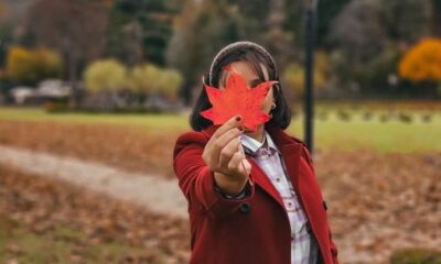 A woman in a red coat holds a vibrant maple leaf in a scenic park during fall.