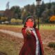 A woman in a red coat holds a vibrant maple leaf in a scenic park during fall.