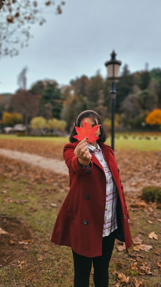 A woman in a red coat holds a vibrant maple leaf in a scenic park during fall.