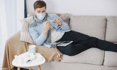 Man wearing a mask checks a thermometer while resting on a couch, surrounded by tissues.