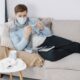 Man wearing a mask checks a thermometer while resting on a couch, surrounded by tissues.