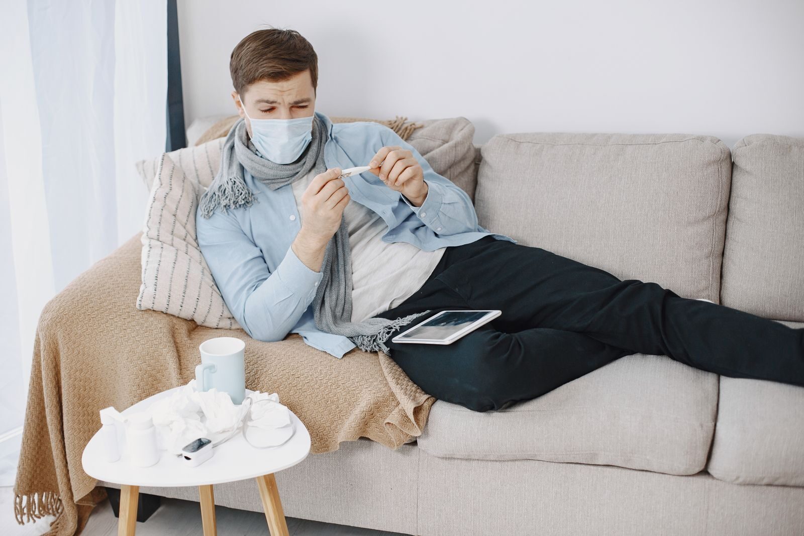 Man wearing a mask checks a thermometer while resting on a couch, surrounded by tissues.