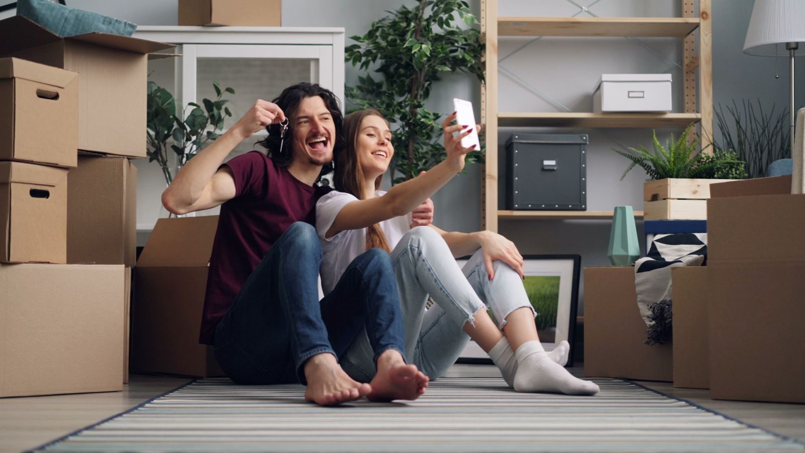 A joyful couple sitting amidst boxes, taking a selfie in their new home.